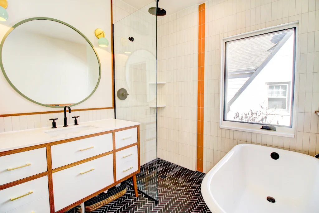 Cherry St. bathroom remodel by Northbuilt Construction, featuring a freestanding soaking tub, custom wood vanity, black herringbone tile flooring, vertical cream wall tile with a bold orange accent strip, and matte black fixtures for a modern, mid-century look.