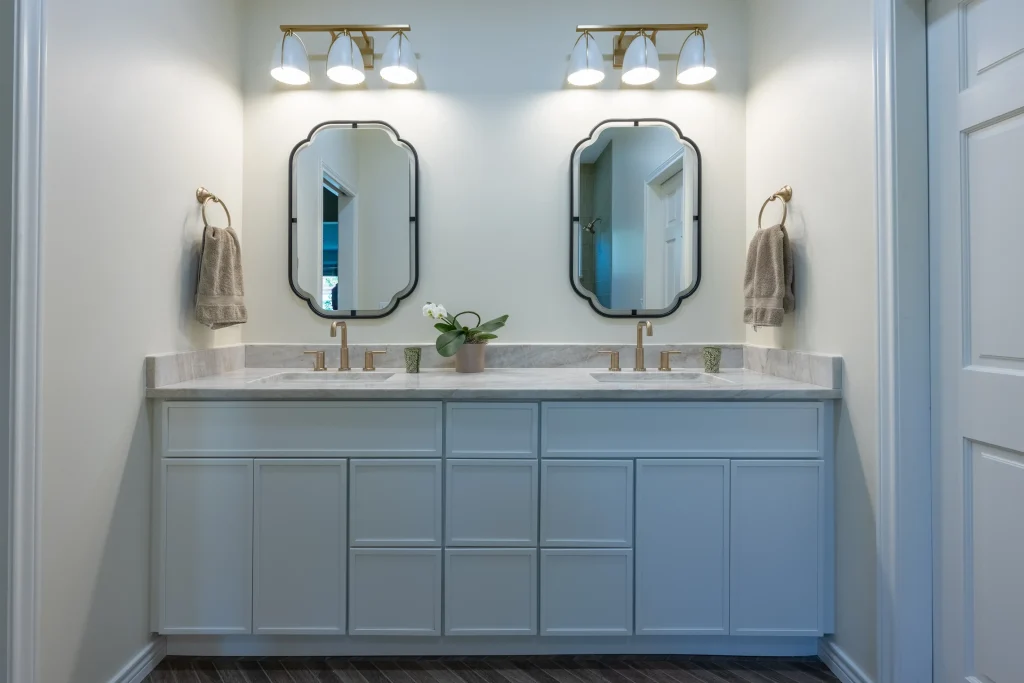 Modern bathroom remodeling in Denver, CO featuring a white double vanity with marble countertops, stylish gold fixtures, black-framed decorative mirrors, and contemporary lighting.