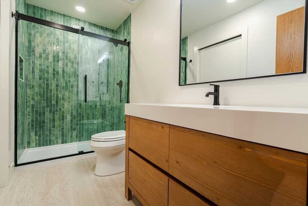 Modern bathroom with glass shower, green tiled walls, white toilet, and a wooden vanity with a black faucet.